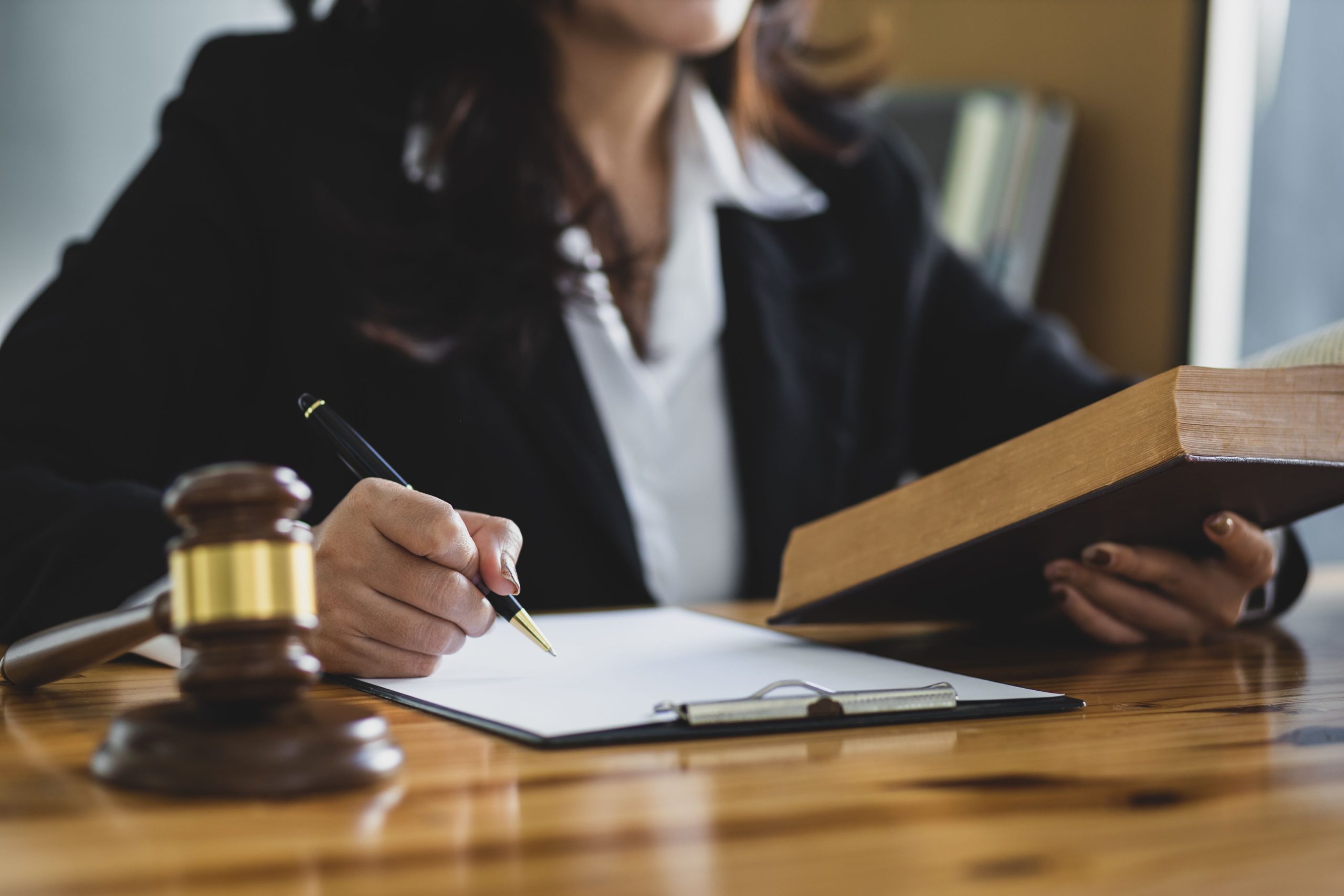 Woman in professional attire sitting at a wooden desk, writing on a clipboard with a pen in one hand and holding a large book in the other. A judge's gavel is placed on the desk in the foreground.