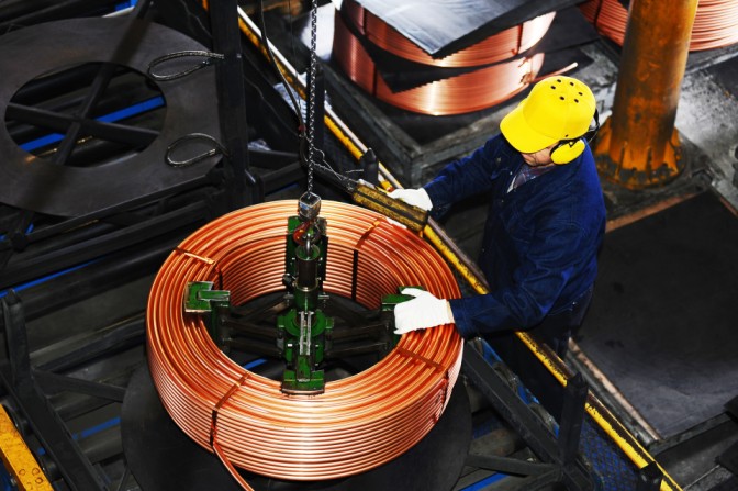 Worker with safety equipment inspecting copper wire coil suspended by chain in factory warehouse